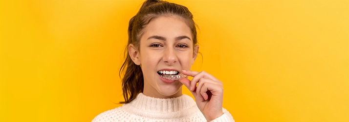 Cheerful young woman on yellow background showing an invisible tooth aligner in her hand.